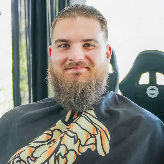 Front portrait of a smiling client showing off his beard trim and slicked-back hairstyle wearing a barber cape.